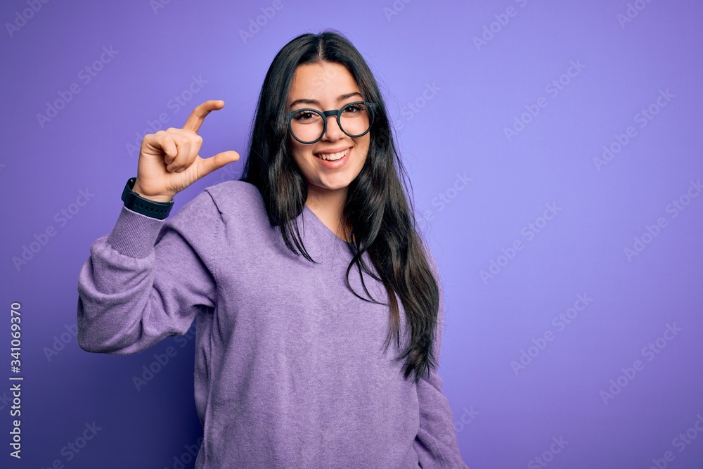 Young brunette woman wearing glasses over purple isolated background smiling and confident gesturing with hand doing small size sign with fingers looking and the camera. Measure concept. Young brunette woman wearing glasses over purple isolated background smiling and confident gesturing with hand doing small size sign with fingers looking and the camera. Measure concept.