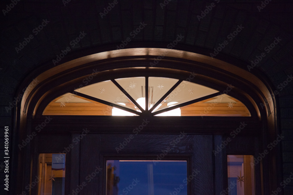 Beautiful illuminated brown wood arched window at twilight with a chandelier indoors. Front door illuminated arch window.
