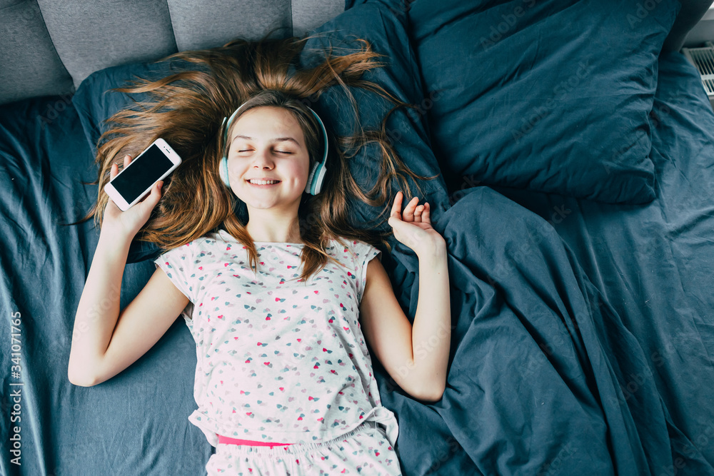 Fototapeta premium .Caucasian beautiful teenager girl with long hair in pajamas lying in bed with his eyes closed and enjoying cool music in modern headphones and holding a phone. Typical teenager morning..bedding blue