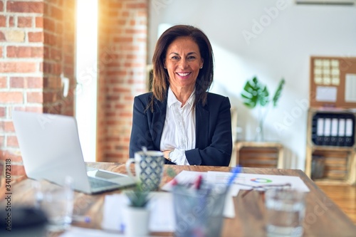 Middle age beautiful businesswoman working using laptop at the office happy face smiling with crossed arms looking at the camera. Positive person.