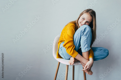 teenage girl looks at side and.sitting on white modern chair with legs up. Beautiful girl in yellow sweater and blue jeans in home interior. Trendy casual outfit. place for text