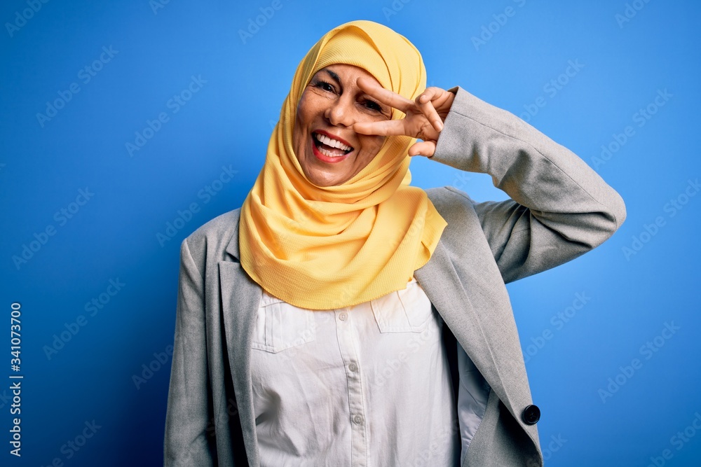 Middle age brunette business woman wearing muslim traditional hijab over blue background Doing peace symbol with fingers over face, smiling cheerful showing victory Middle age brunette business woman wearing muslim traditional hijab over blue background Doing peace symbol with fingers over face, smiling cheerful showing victory