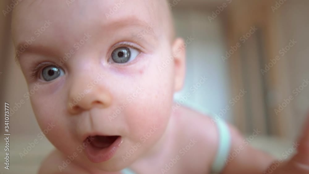 Close up of baby looking straight at the camera and trying to touch it. Big grey eyes. Surprised face expression of a toddler, mouth open. Family lifestile. Bedroom. Girl infant is lying on the bed. Close up of baby looking straight at the camera and trying to touch it. Big grey eyes. Surprised face expression of a toddler, mouth open. Family lifestile. Bedroom. Girl infant is lying on the bed.
