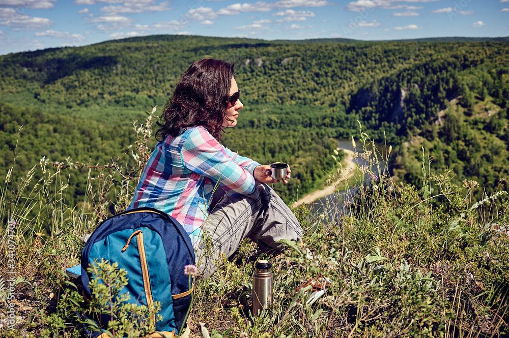 Young happy woman with a backpack sits on a cliff and looks at the valley below Young happy woman with a backpack sits on a cliff and looks at the valley below