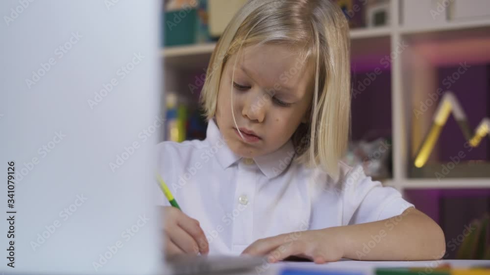 Distance learning due quarantine, online education. Long blond hair preschooler boy in white shirt watching something on the laptop device at the desk in the cozy kid's room. Distance learning due quarantine, online education. Long blond hair preschooler boy in white shirt watching something on the laptop device at the desk in the cozy kid's room.