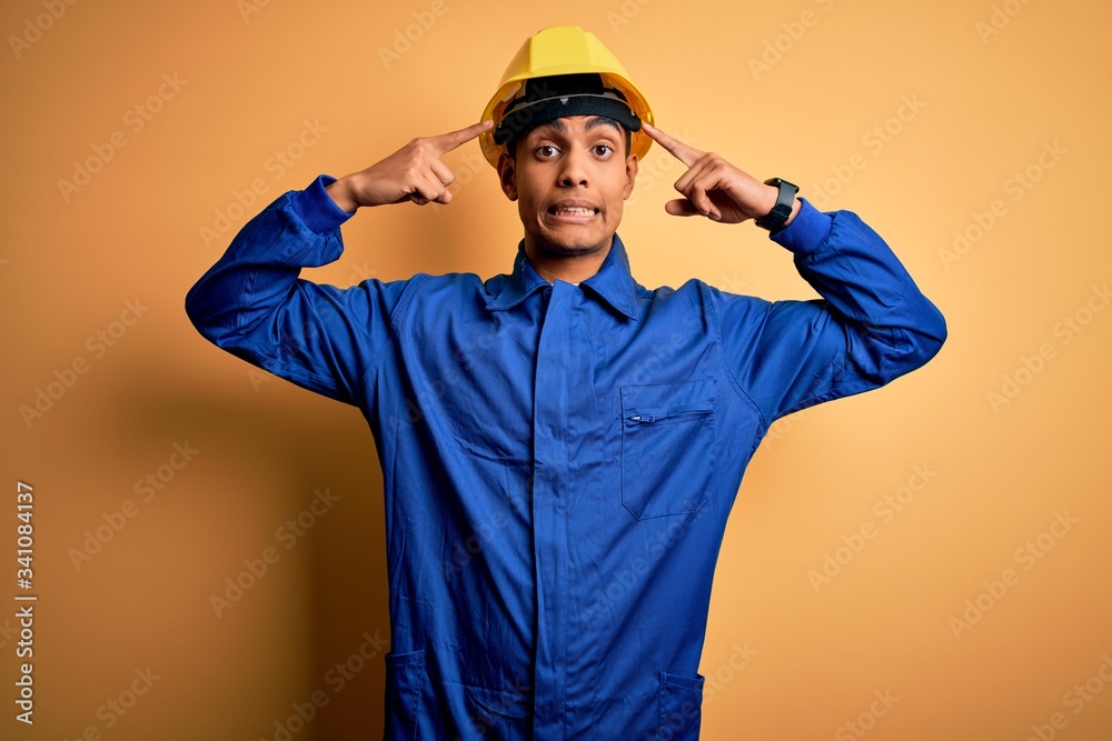 Young handsome african american worker man wearing blue uniform and security helmet smiling pointing to head with both hands finger, great idea or thought, good memory