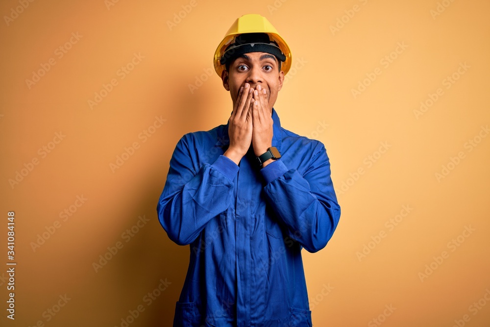 Young handsome african american worker man wearing blue uniform and security helmet laughing and embarrassed giggle covering mouth with hands, gossip and scandal concept Young handsome african american worker man wearing blue uniform and security helmet laughing and embarrassed giggle covering mouth with hands, gossip and scandal concept