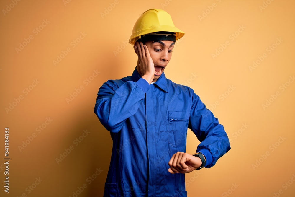 Young handsome african american worker man wearing blue uniform and security helmet Looking at the watch time worried, afraid of getting late
