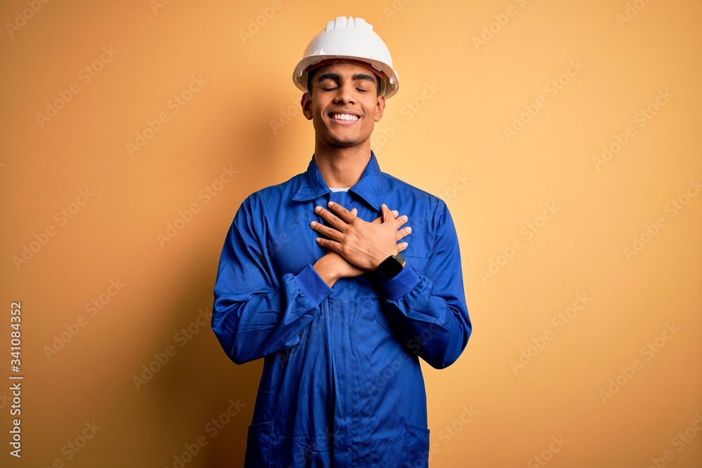 Young handsome african american worker man wearing blue uniform and security helmet smiling with hands on chest with closed eyes and grateful gesture on face. Health concept.