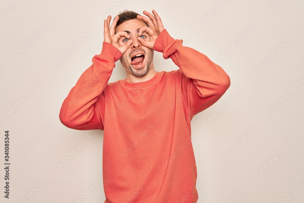 Young handsome man with blue eyes wearing casual sweater standing over white background doing ok gesture like binoculars sticking tongue out, eyes looking through fingers. Crazy expression. Young handsome man with blue eyes wearing casual sweater standing over white background doing ok gesture like binoculars sticking tongue out, eyes looking through fingers. Crazy expression.