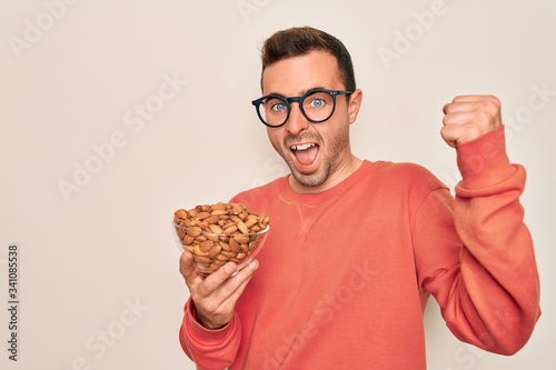 Handsome man with blue eyes holding bowl with healthy almonds snack over white background screaming proud and celebrating victory and success very excited  cheering emotion