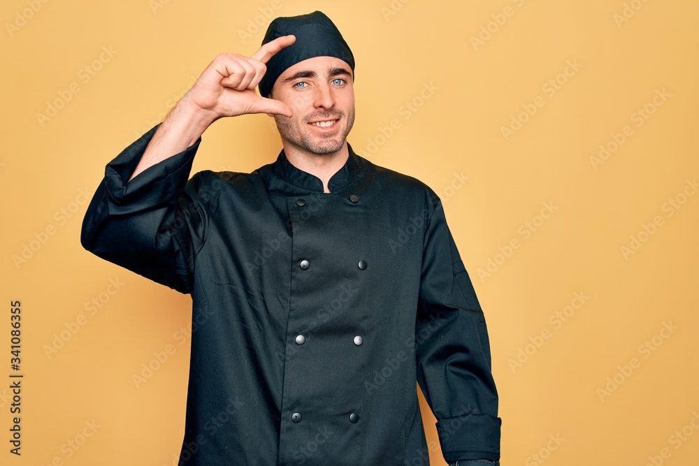 Young handsome cooker man with blue eyes wearing uniform and hat over yellow background smiling and confident gesturing with hand doing small size sign with fingers looking and the camera. Measure