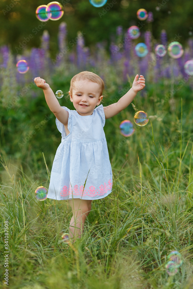 Cute little girl in blue dress try to catch soap bubbles. Smiling baby