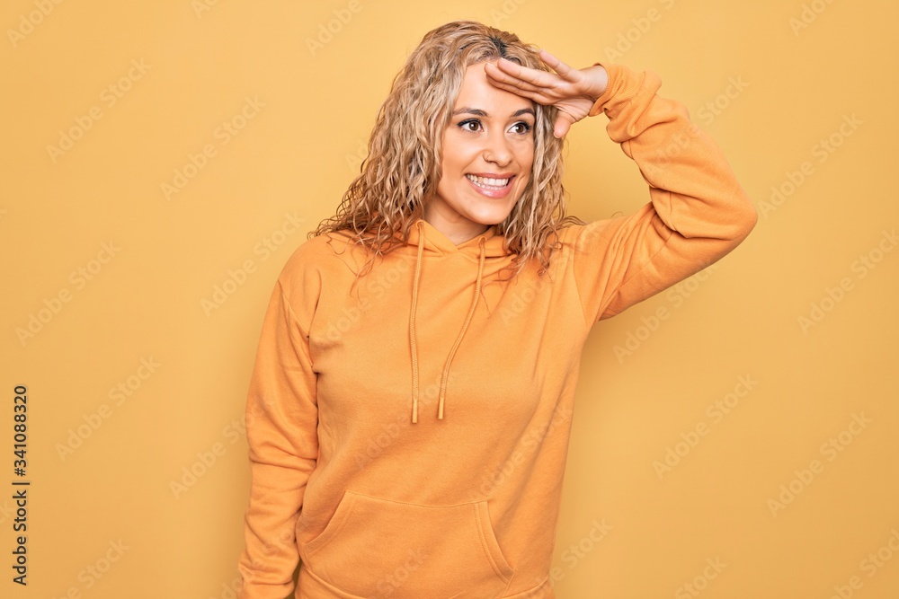 Young beautiful blonde sporty woman wearing casual sweatshirt over yellow background very happy and smiling looking far away with hand over head. Searching concept. Young beautiful blonde sporty woman wearing casual sweatshirt over yellow background very happy and smiling looking far away with hand over head. Searching concept.