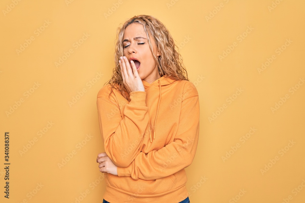 Young beautiful blonde sporty woman wearing casual sweatshirt over yellow background bored yawning tired covering mouth with hand. Restless and sleepiness. Young beautiful blonde sporty woman wearing casual sweatshirt over yellow background bored yawning tired covering mouth with hand. Restless and sleepiness.