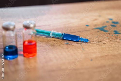 Close-up of a table in a laboratory on which there are two drugs - vaccines - blue and red color, next to which is a syringe and a blue spilled liquid.
