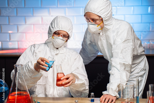 Two young female chemists are experimenting in a laboratory to invent a new vital vaccine. Wearing a protective suit, goggles and a masks, pouring the liquid from the flask and from the syringe