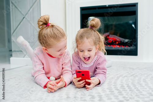 Two young focused children playing a smartphone with no name lying on the living room floor. Young children and technology, sisters play with a mobile phone, watch videos or play games photo