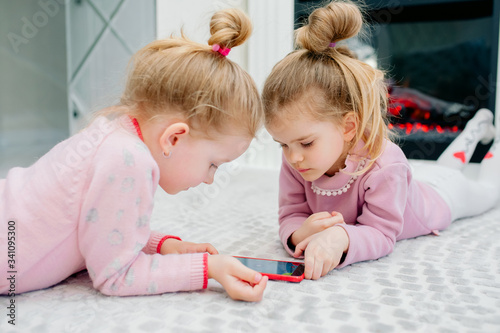 Two young focused children playing a smartphone with no name lying on the living room floor. Young children and technology, sisters play with a mobile phone, watch videos or play games
