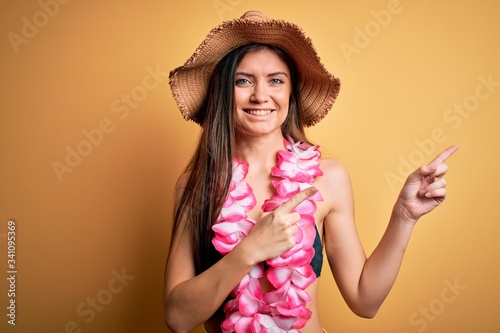 Young beautiful woman with blue eyes on vacation wearing bikini and hawaiian lei smiling and looking at the camera pointing with two hands and fingers to the side.