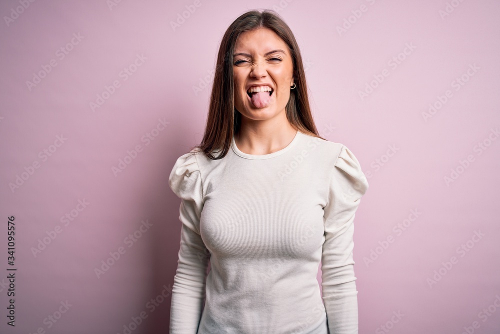 Young beautiful woman with blue eyes wearing casual white t-shirt over pink background sticking tongue out happy with funny expression. Emotion concept. Young beautiful woman with blue eyes wearing casual white t-shirt over pink background sticking tongue out happy with funny expression. Emotion concept.
