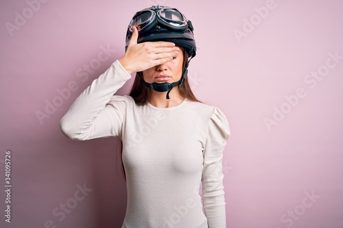 Young beautiful motorcyclist woman with blue eyes wearing moto helmet over pink background covering eyes with hand, looking serious and sad. Sightless, hiding and rejection concept