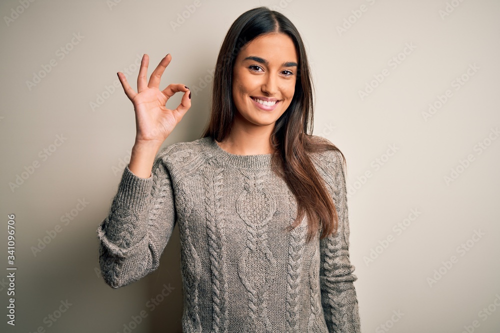 Young beautiful brunette woman wearing casual sweater over isolated white background smiling positive doing ok sign with hand and fingers. Successful expression. Young beautiful brunette woman wearing casual sweater over isolated white background smiling positive doing ok sign with hand and fingers. Successful expression.