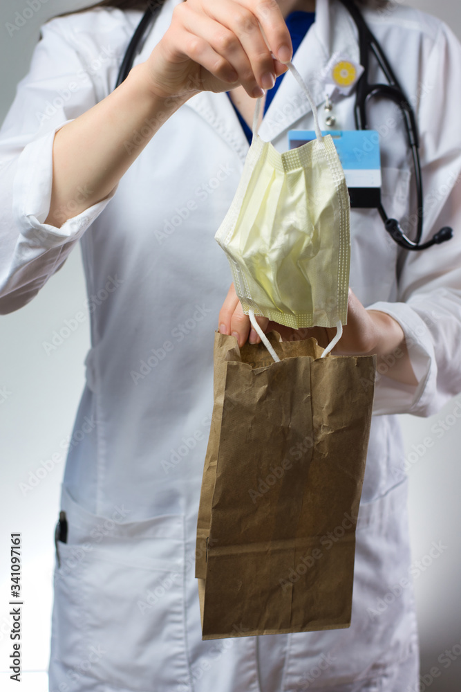 Female healthcare worker demonstrating bagging of mask for coronavirus safety. Paper bag, stethoscope, white coat, and mask visible. Female healthcare worker demonstrating bagging of mask for coronavirus safety. Paper bag, stethoscope, white coat, and mask visible.