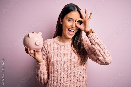 Young beautiful brunette woman holding piggy bank saving money for retirement with happy face smiling doing ok sign with hand on eye looking through fingers photo