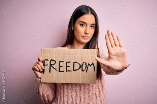 Young beautiful brunette activist woman protesting for freedom holding poster with open hand doing stop sign with serious and confident expression, defense gesture