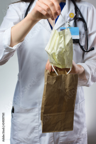 Female healthcare worker demonstrating bagging of mask for coronavirus safety. Paper bag, stethoscope, white coat, and mask visible.