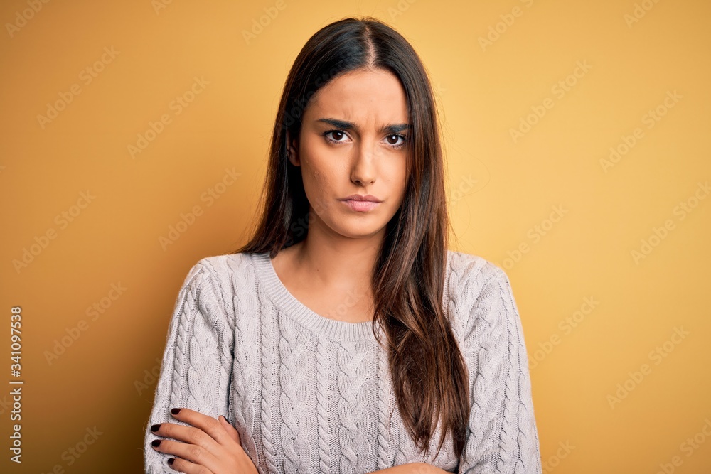 Young beautiful brunette woman wearing white casual sweater over yellow background skeptic and nervous, disapproving expression on face with crossed arms. Negative person. Young beautiful brunette woman wearing white casual sweater over yellow background skeptic and nervous, disapproving expression on face with crossed arms. Negative person.