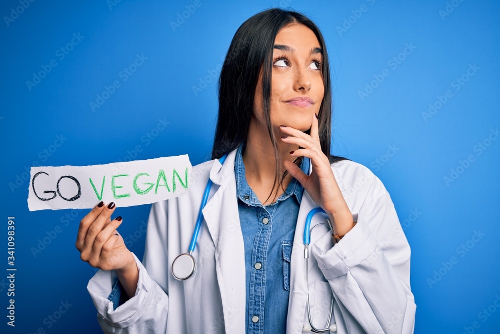 Young beautiful brunette doctor woman wearing coat holding paper with go vegan message serious face thinking about question, very confused idea Young beautiful brunette doctor woman wearing coat holding paper with go vegan message serious face thinking about question, very confused idea