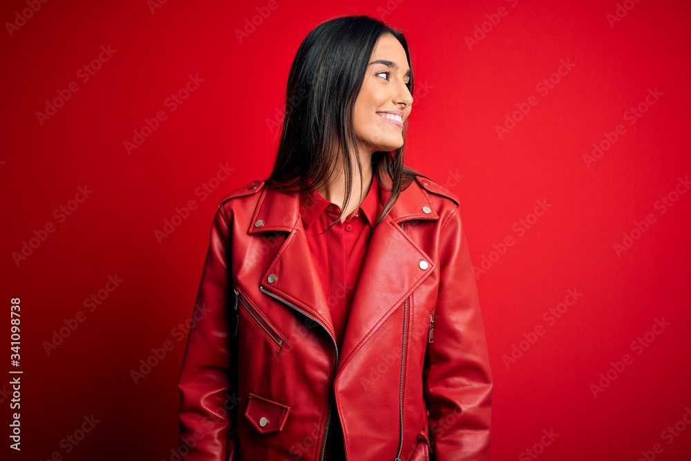 Young beautiful brunette woman wearing casual jacket standing over isolated red background looking away to side with smile on face, natural expression. Laughing confident.