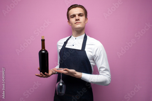 young guy sommelier holds a bottle of red wine on a colored background, an expert on wine in a white shirt and apron, the waiter offers wine photo
