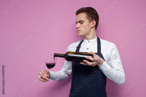 Obraz na płótnie young professional waiter in uniform pours wine into a glass on a pink backgroun