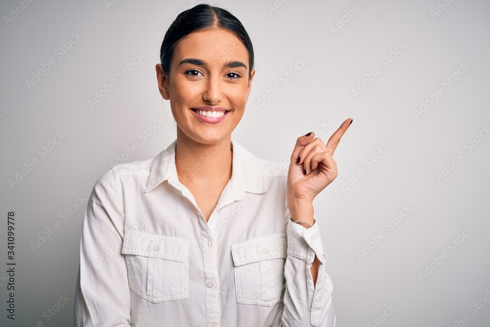 Young beautiful brunette woman wearing casual shirt over isolated white background with a big smile on face, pointing with hand and finger to the side looking at the camera. Young beautiful brunette woman wearing casual shirt over isolated white background with a big smile on face, pointing with hand and finger to the side looking at the camera.