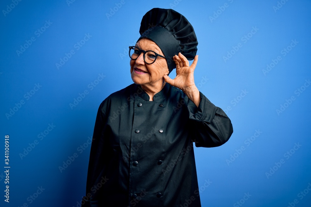 Senior beautiful grey-haired chef woman wearing cooker uniform and hat over blue background smiling with hand over ear listening an hearing to rumor or gossip. Deafness concept. Senior beautiful grey-haired chef woman wearing cooker uniform and hat over blue background smiling with hand over ear listening an hearing to rumor or gossip. Deafness concept.