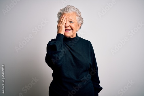 Senior beautiful woman wearing casual black sweater standing over isolated white background covering one eye with hand, confident smile on face and surprise emotion.
