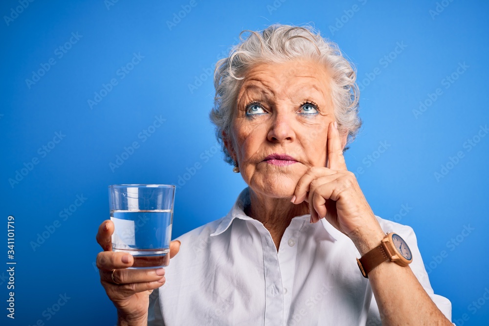 Senior beautiful woman drinking glass of water standing over isolated blue background serious face thinking about question, very confused idea Senior beautiful woman drinking glass of water standing over isolated blue background serious face thinking about question, very confused idea