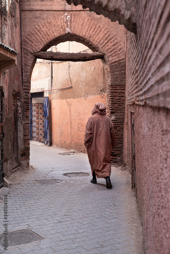 Muslim man with djellaba and kufi strolling alley with shop on the street, blurred background