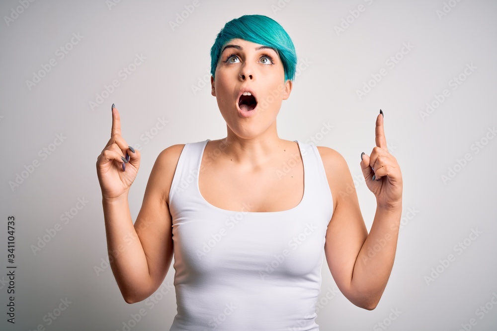Young beautiful woman with blue fashion hair wearing casual t-shirt over white background amazed and surprised looking up and pointing with fingers and raised arms.