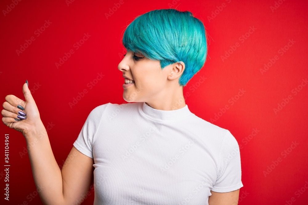Young beautiful woman with blue fashion hair wearing casual t-shirt over red background Looking proud, smiling doing thumbs up gesture to the side Young beautiful woman with blue fashion hair wearing casual t-shirt over red background Looking proud, smiling doing thumbs up gesture to the side