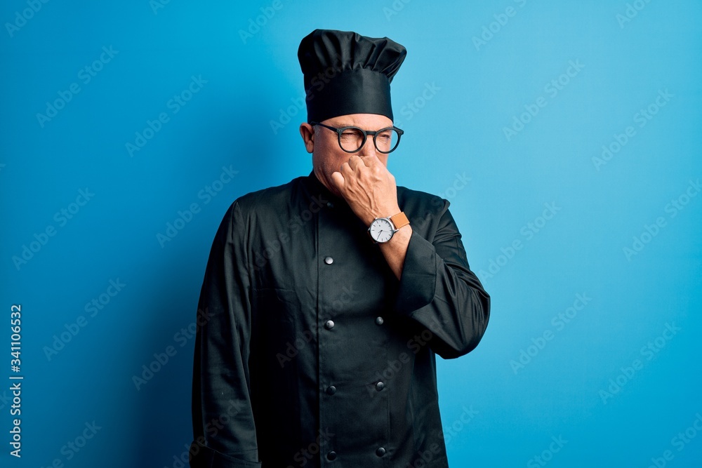 Middle age handsome grey-haired chef man wearing cooker uniform and hat smelling something stinky and disgusting, intolerable smell, holding breath with fingers on nose. Bad smell Middle age handsome grey-haired chef man wearing cooker uniform and hat smelling something stinky and disgusting, intolerable smell, holding breath with fingers on nose. Bad smell