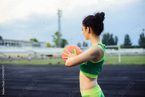 Beautiful curly girl flexing muscles in the stadium. Warm up with the ball. The girl goes in for sports. Light green tracksuit.