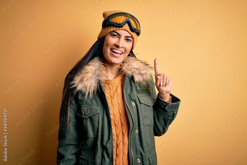 Young beautiful hispanic woman wearing ski glasses and coat for winter weather pointing finger up with successful idea. Exited and happy. Number one.