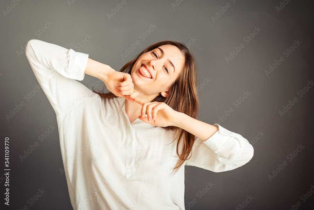 Fototapeta premium Young girl in a white shirt, hands up, on a gray background. Honest and emotional on the banner, billboard, billboard. No retouch. Without make-up.