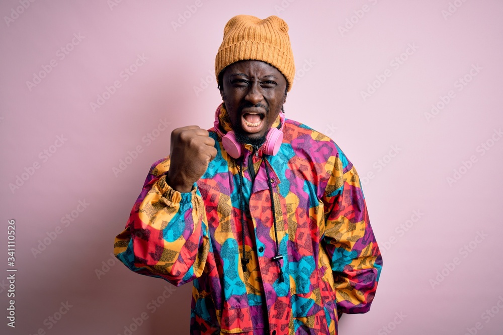 Young handsome african american man wearing colorful coat and cap over pink background angry and mad raising fist frustrated and furious while shouting with anger. Rage and aggressive concept.