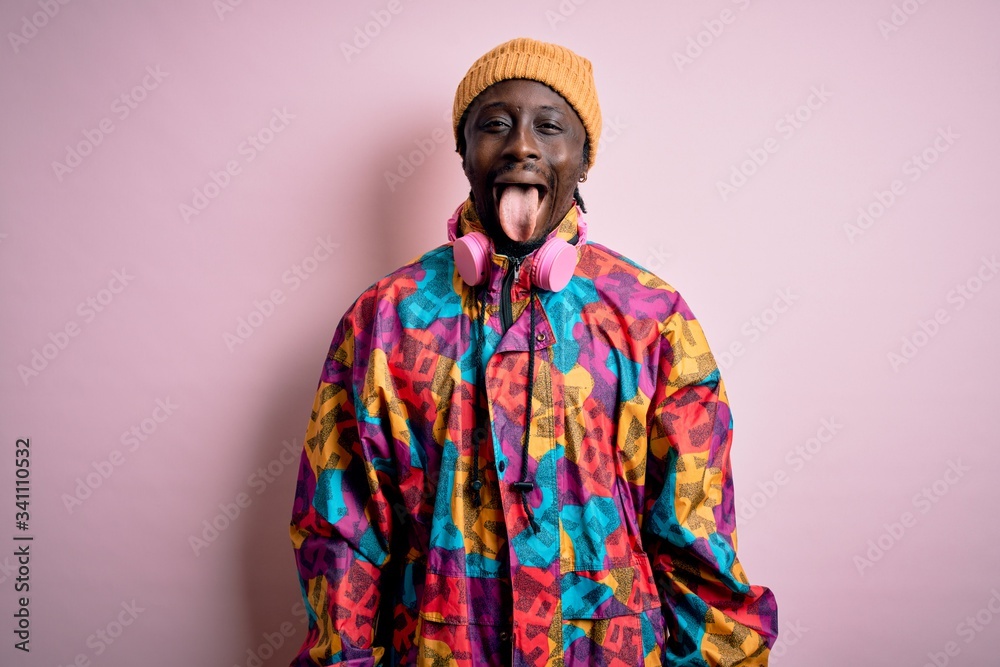 Young handsome african american man wearing colorful coat and cap over pink background sticking tongue out happy with funny expression. Emotion concept.