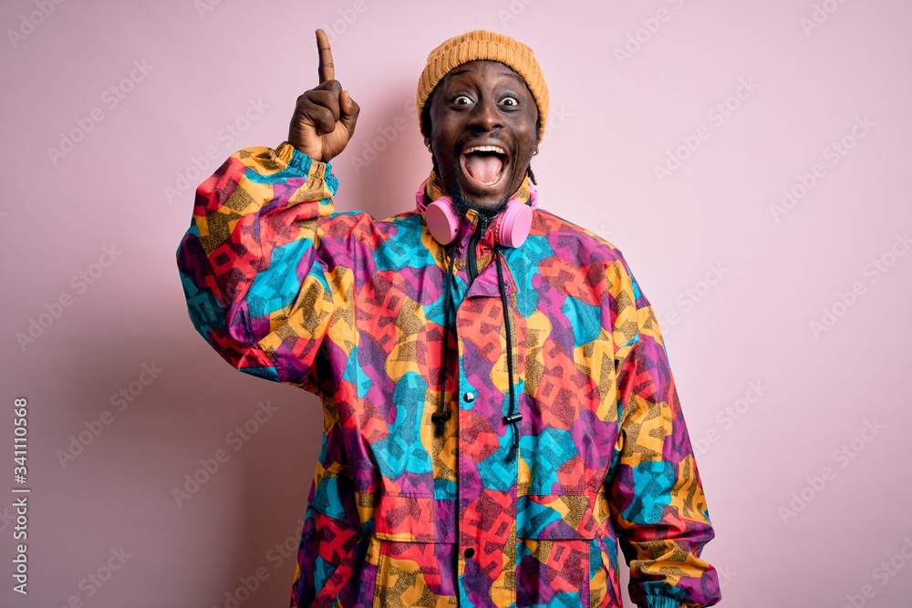 Young handsome african american man wearing colorful coat and cap over pink background pointing finger up with successful idea. Exited and happy. Number one.